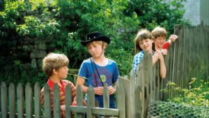 Four children leaning on a weathered wooden fence in a lush garden setting, sunlit and green.