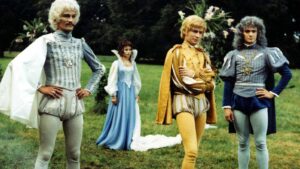Group of four people in elaborate 18th‑century costumes posing in a grassy park setting, with two men in blue-gray outfits, a golden-yellow outfit, and a woman in a light blue gown in the background.