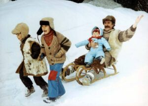 Family of four enjoying a snowy day: two kids lead a man pulling a wooden sled with a baby rider, all in retro winter clothing.