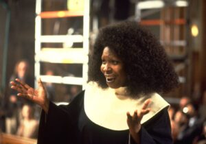 African American woman in a choir robe with a white collar, singing with hands raised and a joyful expression.