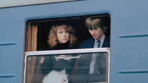 A man and woman in formal clothes sit at a bus window, looking toward each other with neutral expressions.