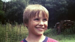 A smiling young boy with blond hair outdoors, grassy field and trees in the background.