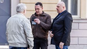 Three men stand outside a building as one man shows a police-style badge or ID card to two others.