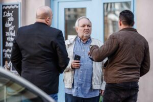 Three men stand on a street, chatting near a storefront with a light blue door; the central man holds a smartphone.