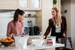 Two women stand at a kitchen island, chatting as they prepare food with bowls, milk, oranges, and utensils on the counter behind them.