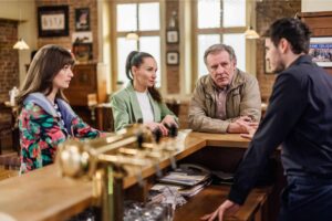 People sit at a wooden bar having a conversation with a bartender; warm, casual cafe/bar atmosphere in the background.