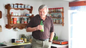 Man with gray hair and mustache in a brown polo holds a green bottle in a cozy kitchen with spice jars and mugs on wooden shelves behind him.