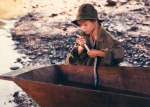 Young boy in a weathered helmet and coat sits in a wooden boat, gripping a rope near a muddy, rocky shore.