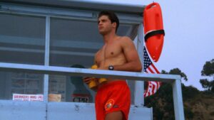Male lifeguard standing shirtless on a station, red trunks, yellow rescue float in hand, watch on wrist, American flag nearby.