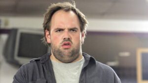 Man indoors with a puzzled, angry expression, gaze straight ahead, wearing a gray shirt and dark jacket.