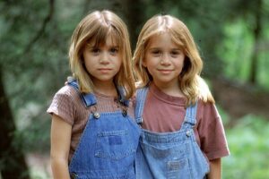 Two smiling young girls with blonde hair in denim overalls posing outdoors in a forested area.