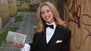 Smiling woman in a black tuxedo with white shirt and bow tie, holding a silver sequined clutch, standing in a graffiti-covered alley.