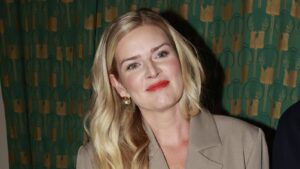 Portrait of a smiling blonde woman wearing a beige blazer and gold earrings, against a green patterned backdrop.