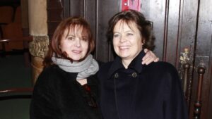 Two women smiling and posing together indoors, one with a gray scarf and black coat, the other in a dark coat with her arm around her friend's shoulder against a wooden door backdrop.