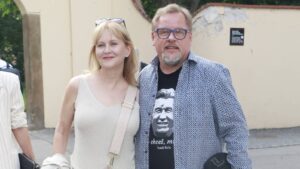 Smiling couple posing outdoors: woman in a beige sleeveless top with a light pink crossbody bag, man in a blue patterned shirt and blue glasses.