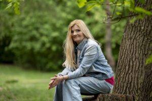 Woman with long blonde hair sits on a log by a tree in a green park, wearing a light denim jacket and jeans.