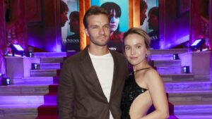 Man and woman posing together on a red-carpet staircase at an event, with colorful posters and purple lighting behind them.