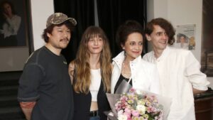 Group of four people posing together, woman in white jacket holds bouquet of pink flowers at center.