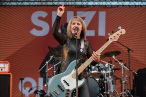 Female rock musician on stage, singing into a microphone while raising one hand and playing a light blue bass guitar.