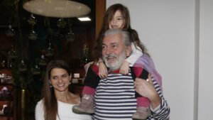 Older man with gray beard carries a smiling child on his shoulders, as two adults stand nearby indoors.