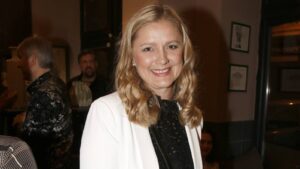 Smiling blonde woman wearing a white blazer over a black lace top at a social event indoors.
