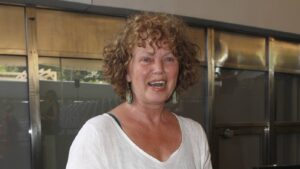 Curly-haired woman in a white blouse with turquoise earrings, smiling at the camera indoors.