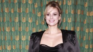 Smiling woman in a black satin dress posing against a teal and gold patterned wall.