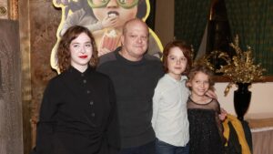 Man posing with three young girls indoors, a large cartoon backdrop behind them showing a popcorn image and oversized glasses.