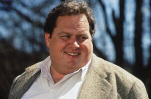 Smiling man outdoors in a beige tweed blazer and light shirt, trees blurred in the background.