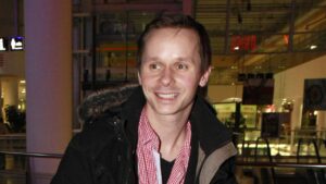 Smiling man in a black jacket with a fur-trimmed hood and red checkered shirt indoors at a shopping mall.