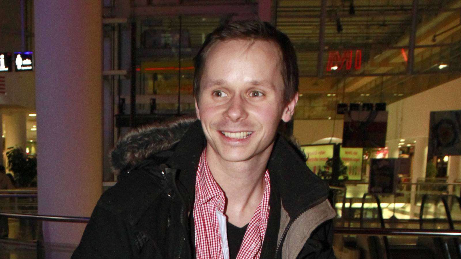 Smiling man in a black jacket with a fur-trimmed hood and red checkered shirt indoors at a shopping mall.