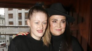 Two women posing closely for a photo indoors, one with light hair and freckles and the other with dark hair wearing a black hat and turtleneck.
