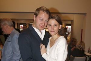Couple posing closely at a social event, smiling at the camera with guests in the background.