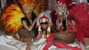Three carnival performers pose together in dazzling feathered costumes, two women in red/gold outfits flanking a man wearing sunglasses and flower leis on a sofa.