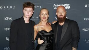 Three adults posing on a red carpet with sponsor logos in the backdrop (left man, center woman, right bearded man).