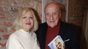 Two smiling older adults stand against a brick wall; the man holds a book with a portrait on the cover.