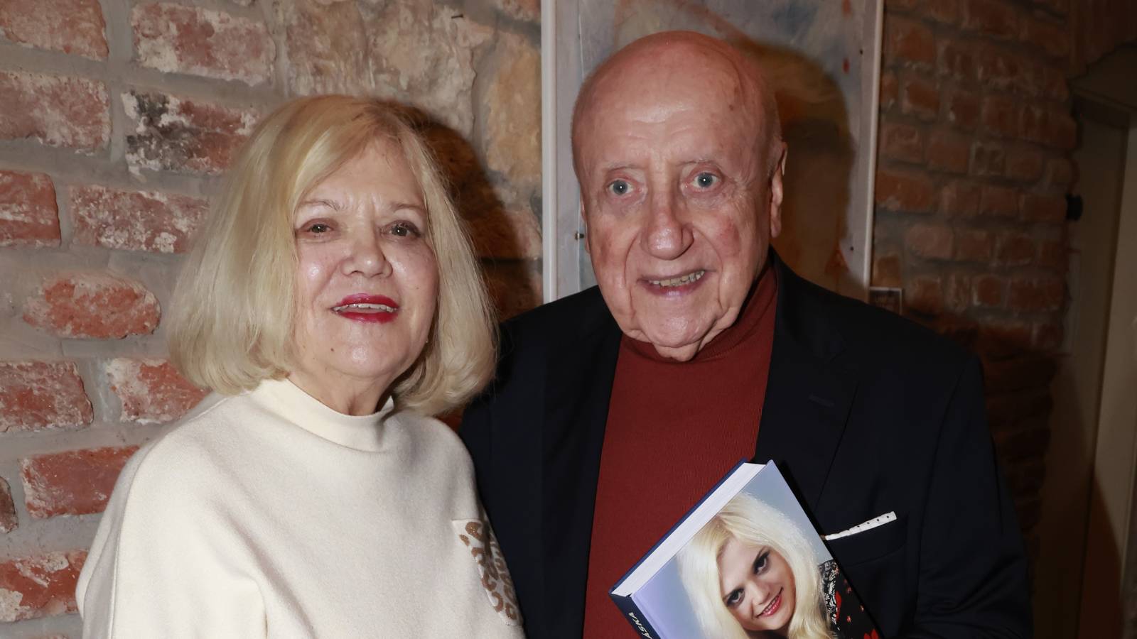 Two smiling older adults stand against a brick wall; the man holds a book with a portrait on the cover.