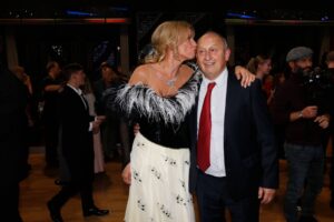 Woman in a black-and-white dress with feather boa kisses an older man in a suit at a gala or party, with photographers in the background.
