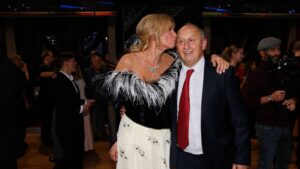 Woman in a black-and-white dress with feather boa kisses an older man in a suit at a gala or party, with photographers in the background.