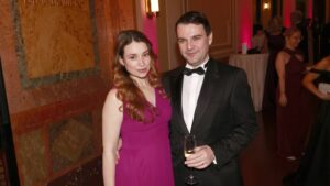Couple in formal attire posing for a photo at a gala; woman in a magenta dress and man in a black tuxedo holding a champagne glass.