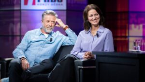 Two smiling hosts sit on a talk-show set with wireless lapel mics clipped to their shirts.