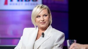 Blonde woman in a white blazer with a lapel microphone, speaking at a panel on a purple-lit stage.
