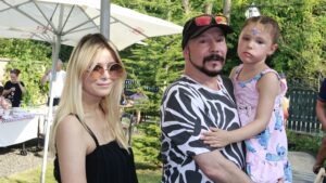 A man in a zebra‑print shirt holds a young girl in a pink dress at an outdoor gathering, with a blonde woman in sunglasses nearby.