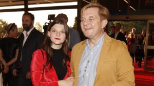 Man in a tan blazer and a young woman in a red jacket pose together on a red carpet at a formal event, smiling at the camera with people in background.