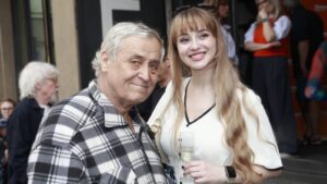 Older man in a plaid shirt posing with a smiling young woman in a white blouse at a social event, holding a wine glass.