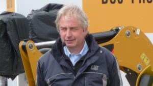 Man wearing a blue CAT jacket with a headset microphone, standing beside a yellow construction machine outdoors.