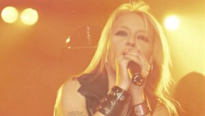 Female singer performing on stage, holding a microphone with warm orange stage lights in the background.