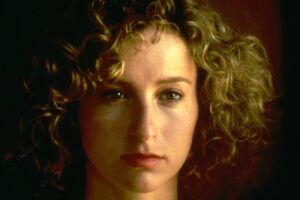 Close-up of a young woman with curly light-brown hair, looking slightly to the left, against a dark background.