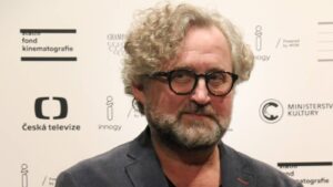 Bearded man with curly gray hair and black glasses posing in front of a sponsor backdrop at an event.
