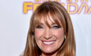 Smiling woman with long light brown hair and bangs, wearing pink lipstick, against a bright event banner background.